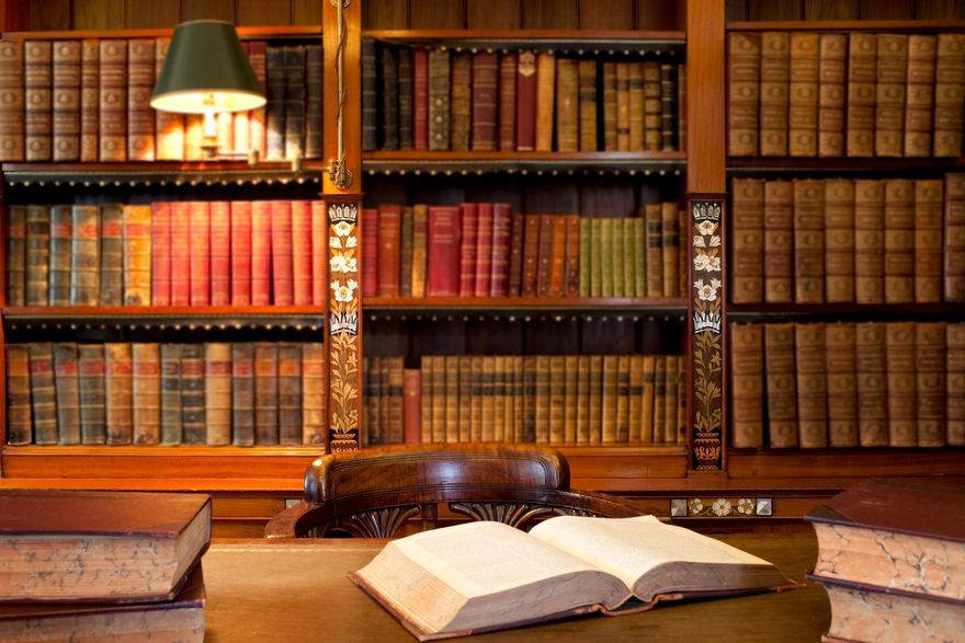 An open book on a desk in front of shelves of leather-bound volumes